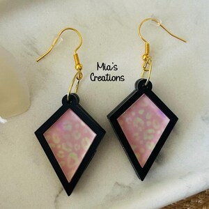 May include: A pair of pink and black diamond-shaped earrings with a gold hook. The earrings have a white and pink abstract pattern.