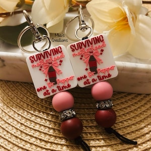 May include: Two keychains with white square pendants featuring the text "SURVIVING motherhood one Dr. Pepper at a time." Each keychain has a pink bow and Dr. Pepper bottle graphic. The keychains have pink and burgundy beads with silver accents.