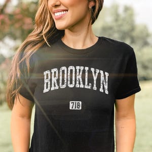 May include: Black crew neck t-shirt featuring the word "BROOKLYN" in white, arched lettering. Below the word "BROOKLYN" is the number "718" in a rectangular box. The shirt is a crew neck style.