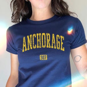 May include: Navy blue crop top with the word "ANCHORAGE" in yellow block letters. Below the word is the number "907" in a yellow rectangle. The shirt has short sleeves.