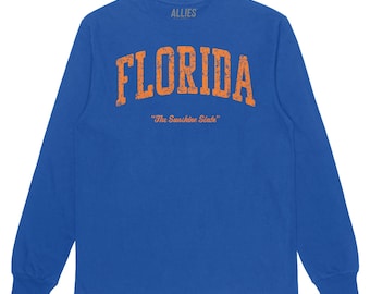 May include: A royal blue long-sleeved t-shirt with an orange vintage-style graphic that reads "FLORIDA" with the text "The Sunshine State" underneath.