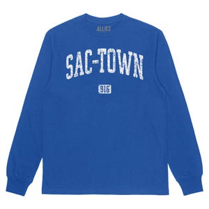 Puede incluir: Una camiseta de manga larga azul real con un gráfico blanco desgastado que dice "SAC-TOWN" con "916" debajo. La camiseta tiene cuello redondo.