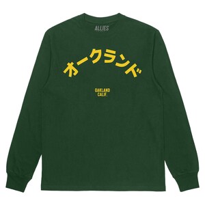 Könnte beinhalten: Ein dunkelgrünes Langarmshirt mit einer gelben Grafik des japanischen Textes "オークランド" (Oakland) in einem Bogen über dem Text "OAKLAND CALIF."
