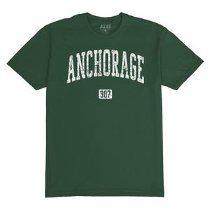 May include: A dark green t-shirt with a white distressed graphic that says "ANCHORAGE 907".