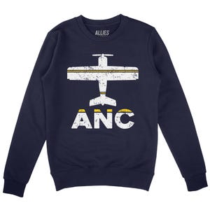 May include: Navy blue sweatshirt featuring a white airplane graphic and the text "ANC" in white with yellow accents. The airplane has a distressed, vintage style.