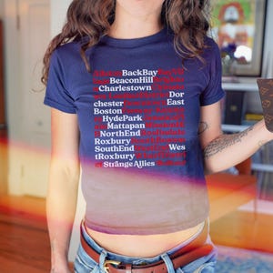May include: A navy blue t-shirt with a gradient effect, displaying a list of city names in red and white. The shirt design includes the text "BackBay", "Beacon Hill", "Charlestown", "Dor", "chester", "East", "Boston", "Hyde Park", "Mattapan", "NorthEnd", "Roxbury", "SouthEnd", "Wes", "tRoxbury", and "Strange Allies".