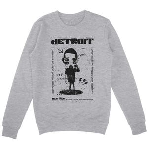 Peut inclure: Un sweat &agrave; manches longues gris avec un imprim&eacute; graphique noir d'un personnage de dessin anim&eacute; avec le mot "DETROIT" au-dessus. Le texte sous le dessin anim&eacute; dit "ESTAMOS TODOS JUNTOS EN ESTO" et "&iquest;POR QUE NO HACER UNA FIESTA?"