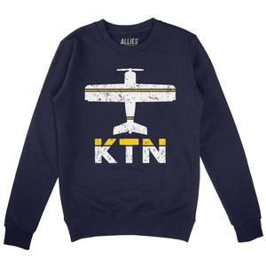 May include: Navy blue sweatshirt featuring a white airplane graphic and the text "KTN" in yellow and white. The airplane has a yellow stripe across the wings and tail.
