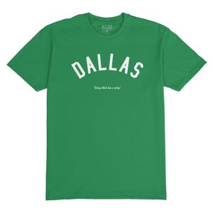 Puede incluir: Camiseta verde con texto blanco que dice "Dallas" en una fuente arqueada. El texto "Every block has a story" est&aacute; impreso en una fuente m&aacute;s peque&ntilde;a debajo de la palabra Dallas.
