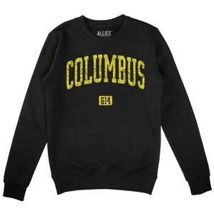 May include: Black crewneck sweatshirt with the word "COLUMBUS" in distressed yellow lettering across the chest. Below the word is the number "614" in a yellow rectangle. The brand name "ALLIES" is printed above the word "COLUMBUS".