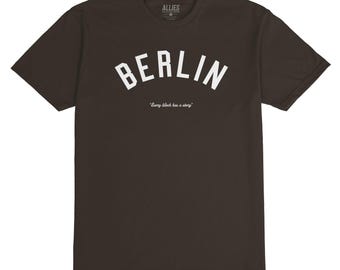 May include: A dark brown t-shirt with a white arch design that says "BERLIN" and the text "Every block has a story".