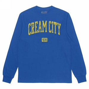 May include: A long-sleeved blue shirt with the text "CREAM CITY" in yellow, arched across the chest. Below the text is the number "414" in a yellow rectangle. The word "ALLIES" is above the main text.