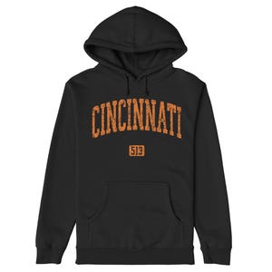 Op de afbeelding: Zwarte hoodie met het woord "CINCINNATI" in oranje, gebogen over de borst. Daaronder staat het nummer "513" in een oranje rechthoek. De sweatshirt heeft een voorzak en een capuchon met trekkoord.