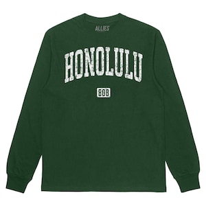 Könnte beinhalten: Ein dunkelgrünes Langarmshirt mit einem weißen Vintage-Grafikdruck, der "HONOLULU" mit "808" darunter zeigt. Das Shirt ist von Allies.