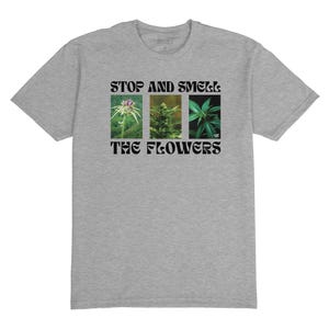 May include: A heather grey t-shirt with the text "Stop and Smell the Flowers" printed in black. The text is in a retro font style. Three images of flowers are displayed in a row below the text.