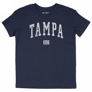 Puede incluir: Camiseta azul marino con la palabra "TAMPA" en letras blancas desgastadas. Debajo, el número "813" también en blanco. La camiseta es de cuello redondo y manga corta.