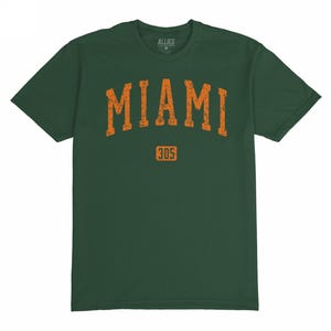 Puede incluir: Camiseta verde oscuro con la palabra "MIAMI" en letras naranja desgastadas en el pecho y el número "305" debajo. La camiseta tiene cuello redondo y mangas cortas. El nombre de la marca "ALLIES" está impreso en la parte superior de la espalda.