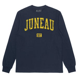 May include: Navy blue long-sleeved t-shirt with a yellow distressed graphic that says "JUNEAU" in a collegiate font. The number "907" is printed in a smaller yellow box below the text. The shirt has a crew neck and long sleeves.