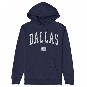 May include: Navy blue hoodie featuring the word "DALLAS" in distressed white lettering. Below the word "DALLAS" is the number "214" in a rectangular box. The sweatshirt has a front pocket and a drawstring hood. A casual garment.