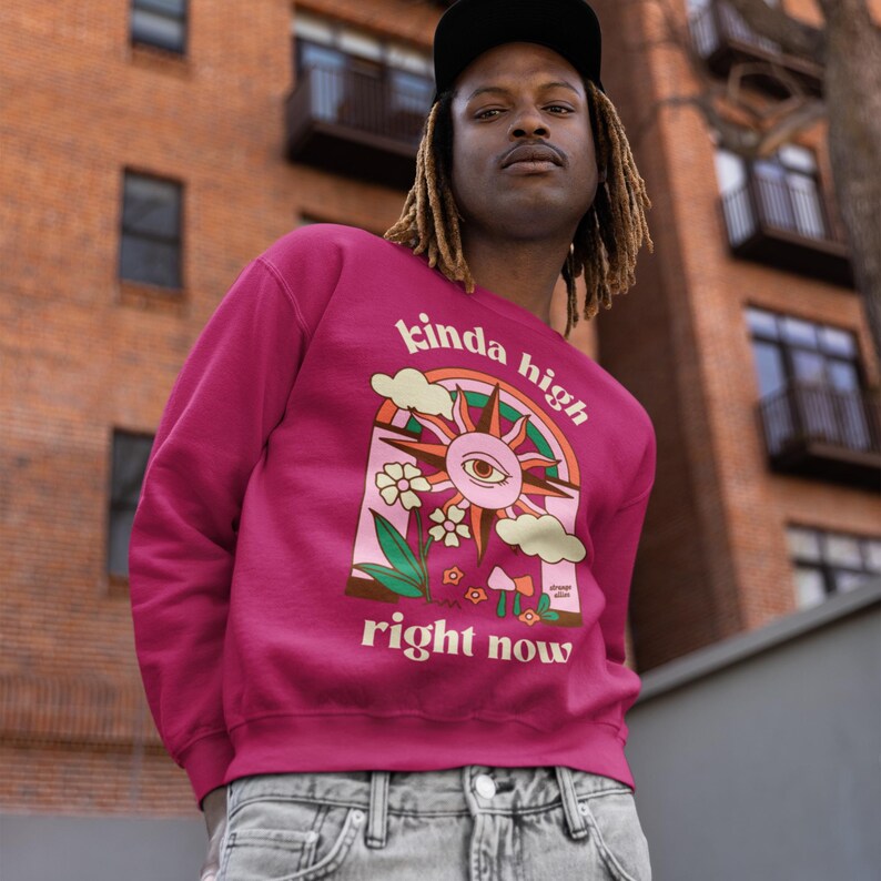 Puede incluir: Una sudadera rosa con un dise&ntilde;o gr&aacute;fico retro que presenta un sol con un ojo, un arco&iacute;ris y el texto "kinda high right now".