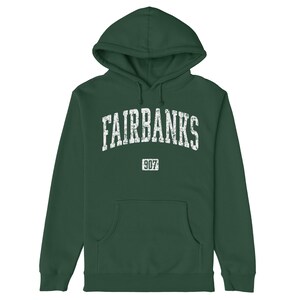 May include: A dark green hoodie featuring the word "FAIRBANKS" in distressed white text across the chest. Below the word is the number "907" in a smaller white box. The sweatshirt has a front pocket and a drawstring hood.