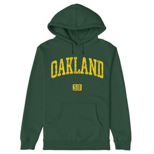 Könnte beinhalten: Ein dunkelgrüner Kapuzenpullover mit einer Vordertasche. Das Wort "OAKLAND" ist in verblichenen gelben Buchstaben auf die Brust gedruckt. Darunter befindet sich die Nummer "510" in einem gelben Rechteck.