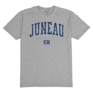 May include: A heather grey t-shirt with a navy blue distressed print of "JUNEAU" in an arch with "907" below it.