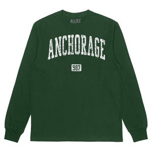 May include: A dark green long-sleeved t-shirt with a white distressed graphic that says "ANCHORAGE" and "907" in a vintage collegiate style.