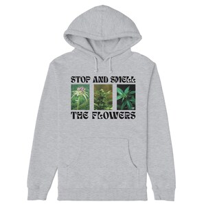 Può includere: Felpa grigia con cappuccio e tasca frontale. La parte anteriore presenta la scritta "STOP AND SMELL THE FLOWERS" sopra tre immagini di piante. Un capo d'abbigliamento casual.