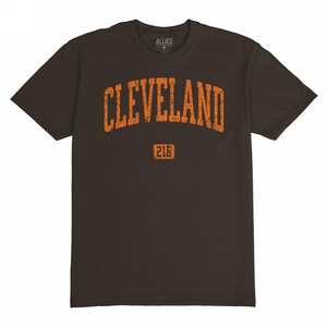 May include: A dark brown t-shirt with the word "CLEVELAND" in distressed orange lettering across the chest. Below the word is the number "216" in a rectangular box. The shirt has short sleeves and a crew neck.