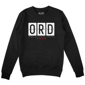 Op de afbeelding: Zwart sweatshirt met een witte afbeelding van een aankomstbord op de luchthaven met "ORD" voor de internationale luchthaven O'Hare in Chicago. De afbeelding bevat ook de coördinaten van de luchthaven en de tekst "ALLIES".