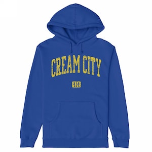 May include: A royal blue hoodie with a front pocket and a hood. The text "CREAM CITY" is printed across the chest in distressed yellow lettering, with "414" below it. The hoodie is made of a soft material.