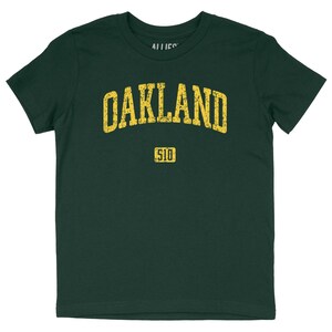 Könnte beinhalten: Dunkelgrünes T-Shirt mit dem Wort "OAKLAND" in verblichenen gelben Buchstaben auf der Brust. Darunter steht die Zahl "510" in Gelb. Das T-Shirt hat einen Rundhalsausschnitt.