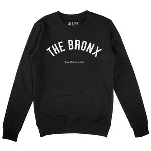 Op de afbeelding: Zwart sweatshirt met witte tekst die "THE BRONX" in een boog vormt. De tekst "Every block has a story" staat onder de boog.