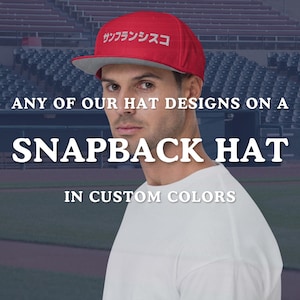 May include: A red snapback hat with a gray brim. The hat has Japanese text on the front. The image also includes the text "ANY OF OUR HAT DESIGNS ON A SNAPBACK HAT IN CUSTOM COLORS."