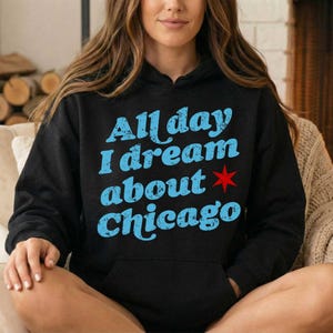 May include: A black hooded sweatshirt featuring the light blue distressed text All day I dream about Chicago with a small red six-pointed star symbol positioned to the right of the word about. The garment includes a front kangaroo pocket.