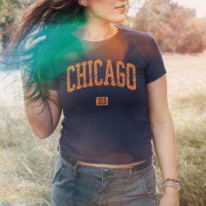 Puede incluir: Camiseta azul marino con la palabra "CHICAGO" en letras naranjas desgastadas. Debajo de la palabra "CHICAGO" está el número "312" en un pequeño rectángulo. La camiseta la lleva una persona en un campo.