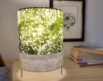 May include: A green and white floral lampshade with a white base and gold metal legs. The lampshade is lit and casting a warm glow.