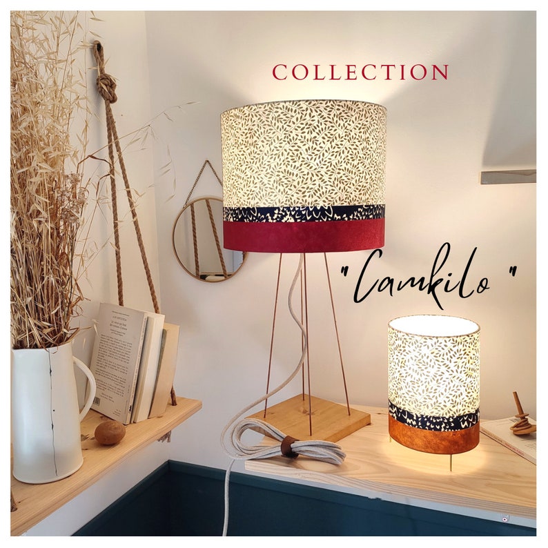 May include: Two table lamps with cylindrical shades featuring a cream leaf pattern, a dark blue band, and a solid base. The taller lamp has a wooden square base and thin copper legs. The text "COLLECTION" and "Camkilo" appear in the image.