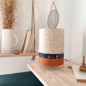 May include: A cylindrical table lamp featuring a top section with a gold leaf pattern on a white background, a middle band of dark blue with a white abstract print, and a solid terracotta orange base, supported by three thin gold metal legs.