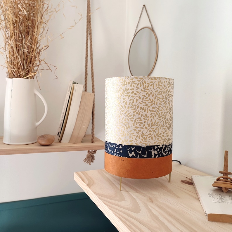 May include: A cylindrical table lamp featuring a top section with a gold leaf pattern on a white background, a middle band of dark blue with a white abstract print, and a solid terracotta orange base, supported by three thin gold metal legs.