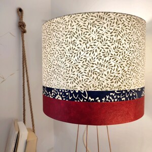 May include: A cylindrical lamp shade featuring a top section with a green leaf pattern on a cream background, a middle band of dark blue with white abstract markings, and a solid deep red bottom section, supported by thin metallic legs.
