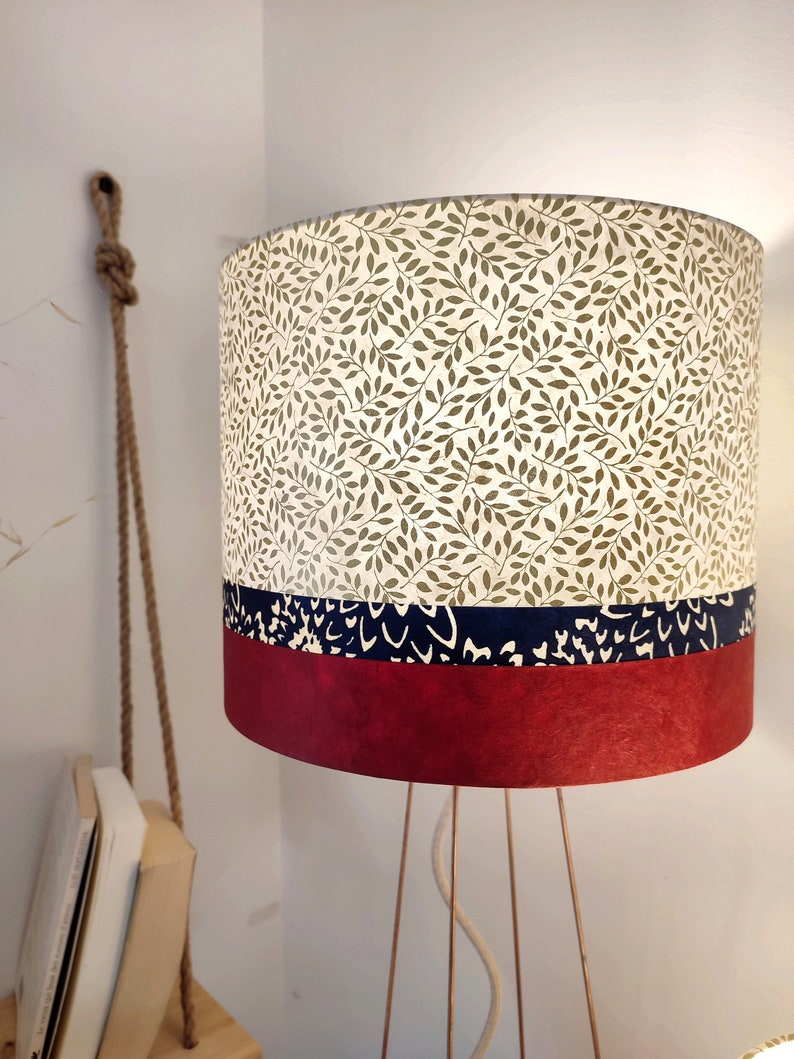 May include: A cylindrical lamp shade featuring a top section with a green leaf pattern on a cream background, a middle band of dark blue with white abstract markings, and a solid deep red bottom section, supported by thin metallic legs.