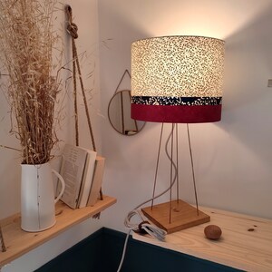 May include: A table lamp with a cylindrical shade featuring a cream and dark green leaf pattern, a thin dark blue band, and a solid maroon base section. The lamp has a square wooden base, thin metal legs, and a light-colored power cord with a plug.