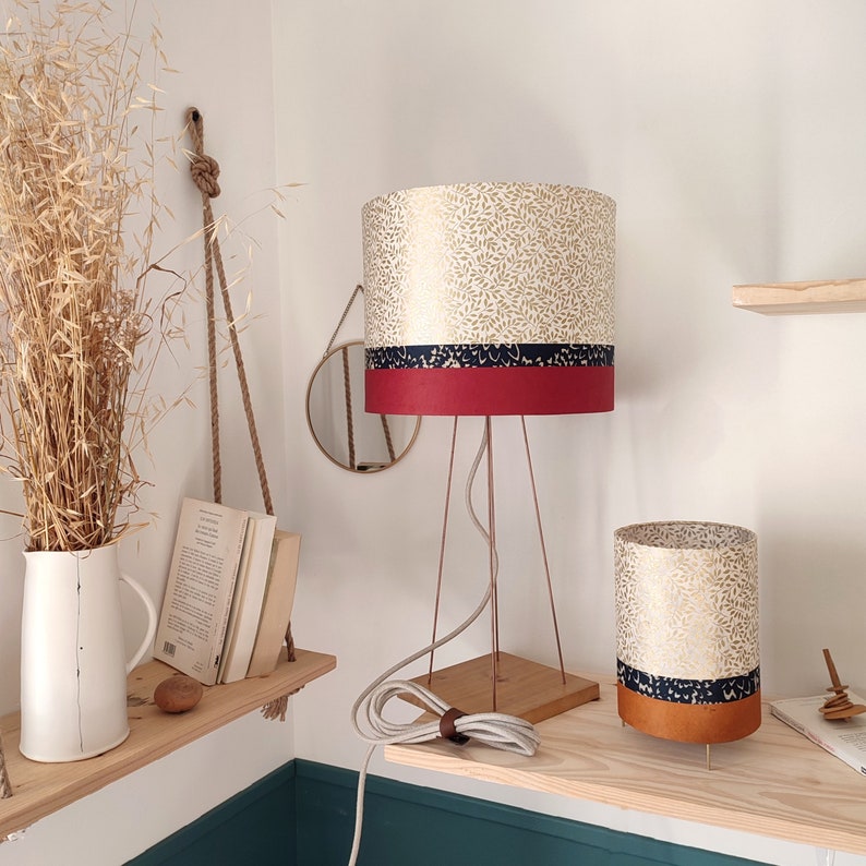 May include: A table lamp with a cylindrical shade featuring a gold leaf pattern, a dark blue band, and a solid red base. The lamp has a thin metal tripod frame and a square wooden base. A matching smaller cylindrical lamp shade sits nearby.