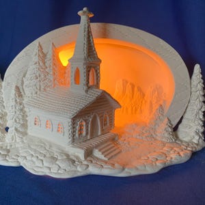 May include: A white ceramic church night light with a steeple, surrounded by trees and a mountain scene. The church has lit windows and a warm orange glow emanating from within. A decorative piece for home decor.