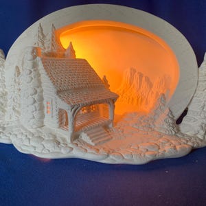 May include: A white ceramic cabin-shaped night light with an illuminated orange background. The cabin has a detailed roof, porch, and chimney, with trees on either side. The light is plugged in with a white cord.