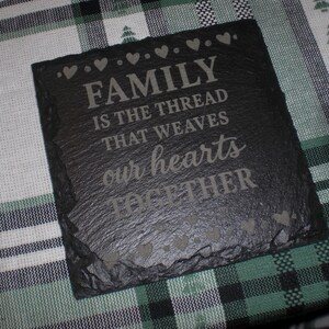 May include: A square, black slate coaster with the engraved text "FAMILY IS THE THREAD THAT WEAVES our hearts TOGETHER". The coaster has a rough, textured surface and is decorated with heart shapes.