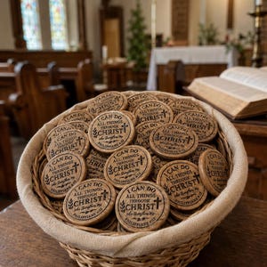 May include: A basket filled with round wooden tokens. Each token has the text "I CAN DO ALL THINGS THROUGH CHRIST" and a cross. The tokens are light brown and the basket is woven.