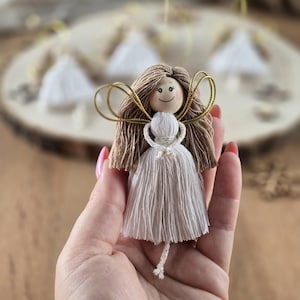 Op de afbeelding: Een witte garen engelversiering met bruin garen haar, een houten hoofd met een geschilderd gezicht en gouden draadvleugels.